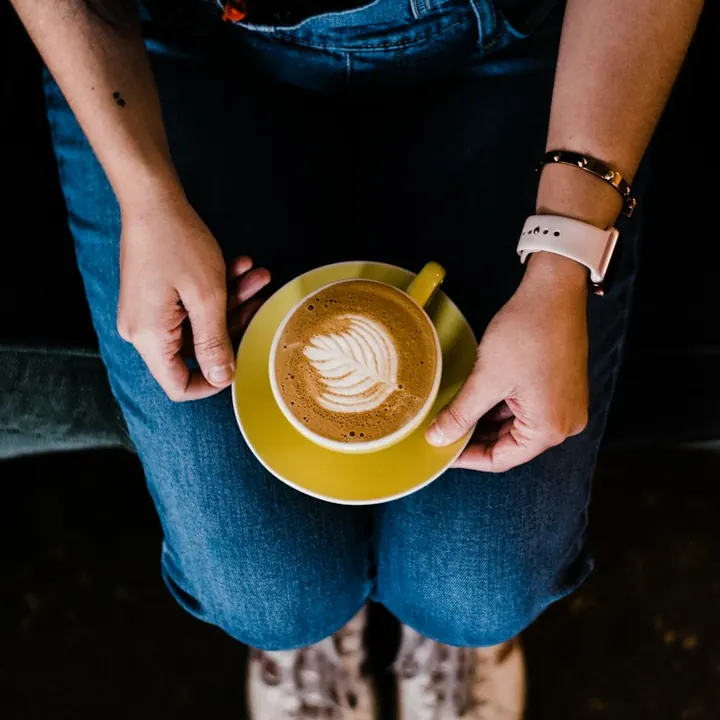 Mãos de uma pessoa sentada no sofá segurando uma xícara de café (imagem ilustrativa)