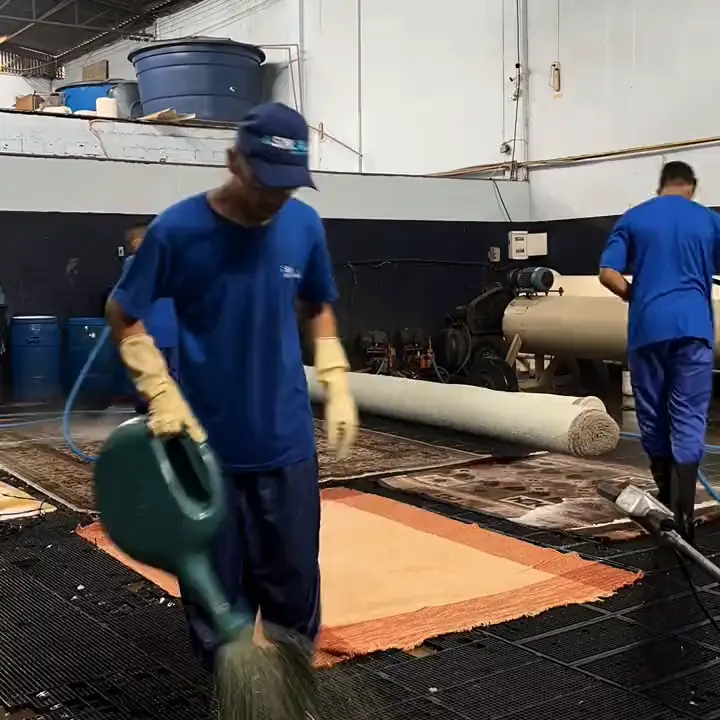 Funcionário da Sinlave aplicando o produto num tapete estendido no piso da lavanderia, com outros tapetes enrolados ao fundo